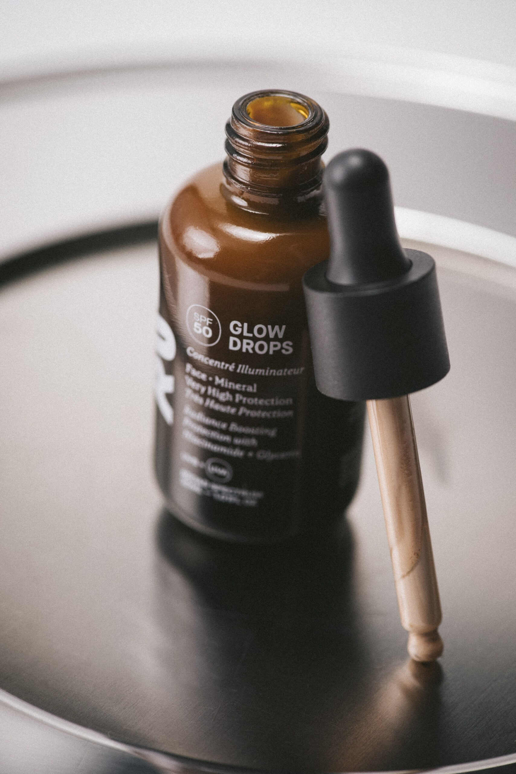 Close-up of a brown glass dropper bottle with a black cap resting in a shallow metal bowl; label reads Glow Drops (skincare product).