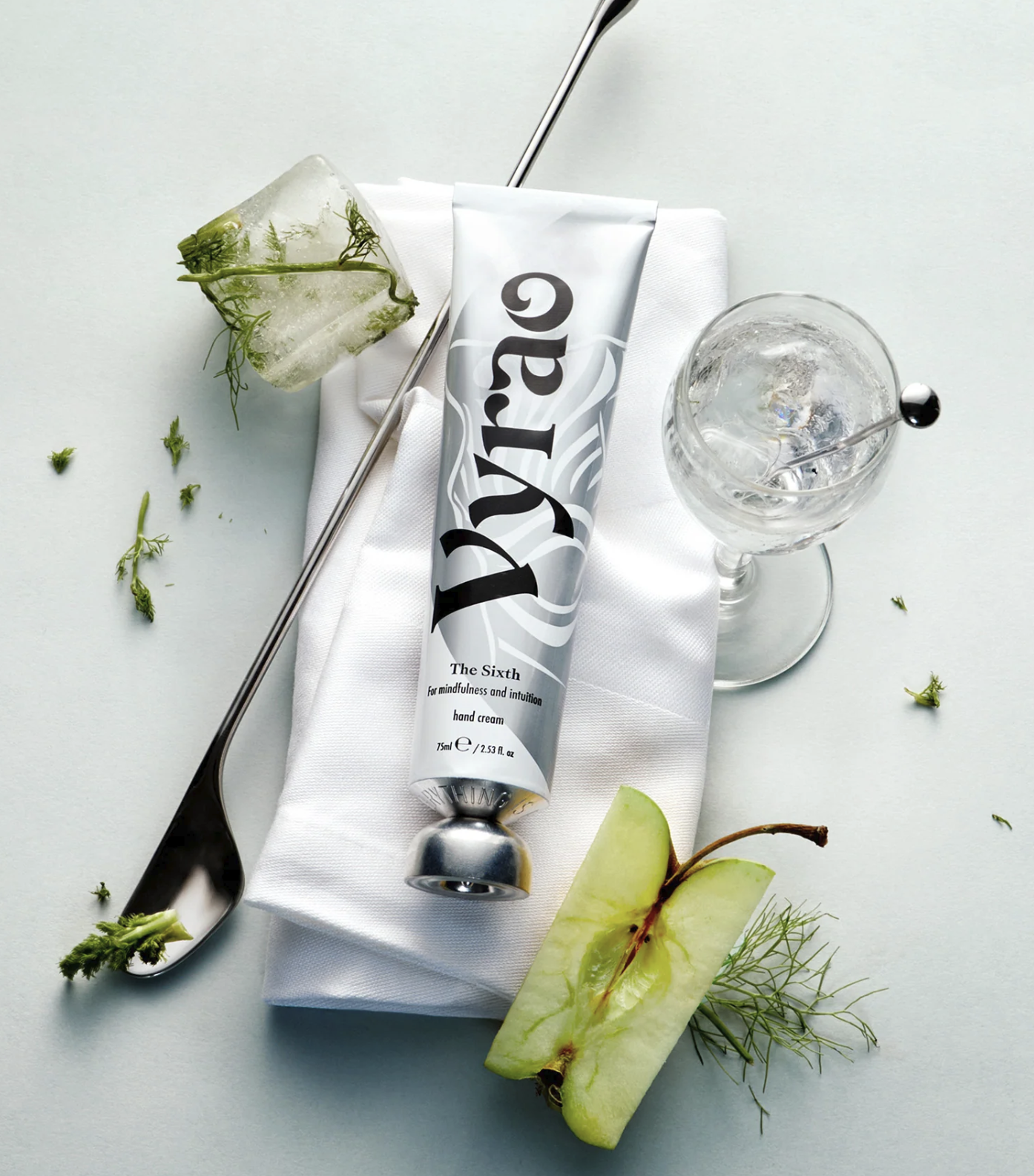 A tube of hand cream labeled 'The Sixth' rests on a white napkin beside a spoon and a glass with ice, with fresh herbs and a green apple slice nearby.