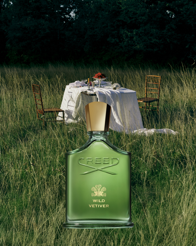 Green Creed Wild Vetiver perfume bottle in the foreground with a rustic outdoor picnic setup in a grassy field behind it.