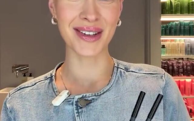 blogg-53943-frame0 - Gimle Parfymeris blogg Victoria smiles at the camera while holding two makeup pens in a denim jacket, with a shelf of colorful beauty products in the background.