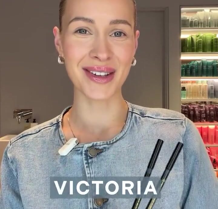 Victoria smiles at the camera while holding two makeup pens in a denim jacket, with a shelf of colorful beauty products in the background.