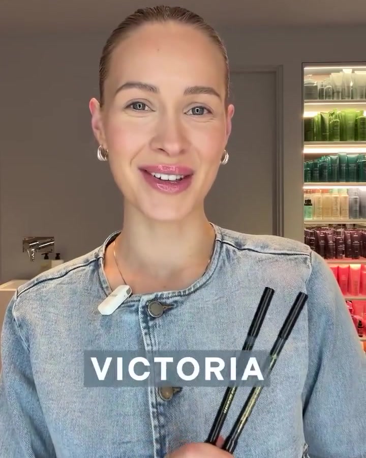 Victoria smiles at the camera while holding two makeup pens in a denim jacket, with a shelf of colorful beauty products in the background.