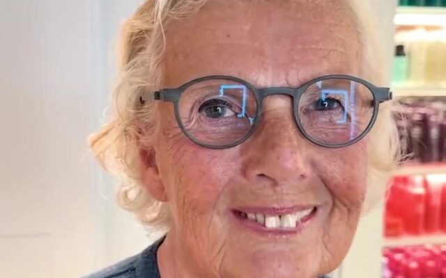 Smiling older woman with round gray glasses in a store, shelves of colorful bottles behind her.]},{