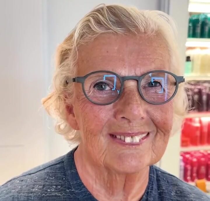 Smiling older woman with round gray glasses in a store, shelves of colorful bottles behind her.]},{