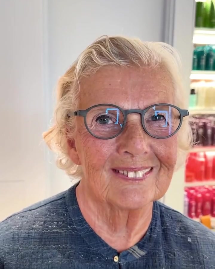 Smiling older woman with round gray glasses in a store, shelves of colorful bottles behind her.]},{