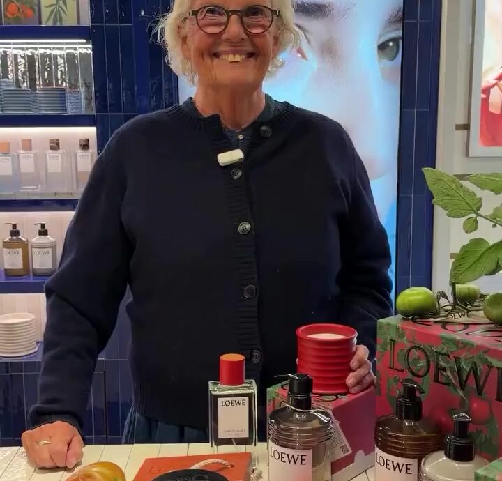 Smiling woman with short white hair and glasses stands behind a Loewe product display at a store counter with bottles and plants besides her.