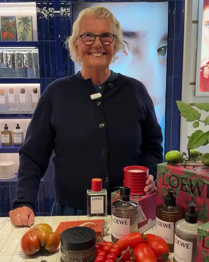 Smiling woman with short white hair and glasses stands behind a Loewe product display at a store counter with bottles and plants besides her.