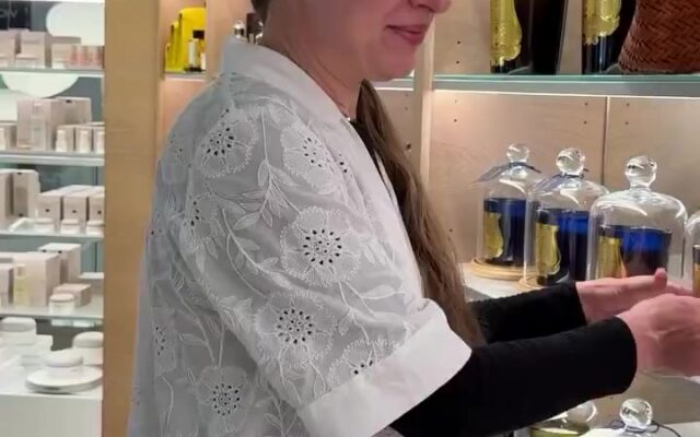 Woman in a white lace top browsing candle display in a store, reaching toward glass cloches on the shelf