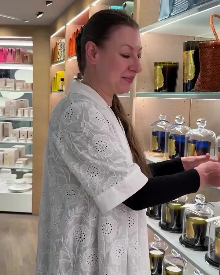 Woman in a white lace top browsing candle display in a store, reaching toward glass cloches on the shelf