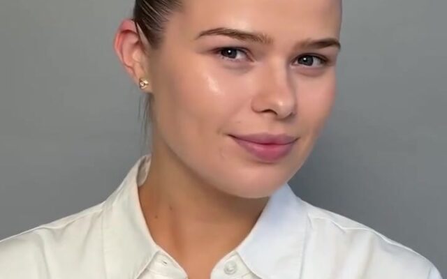 Portrait of a woman with slicked-back hair and a white collared shirt, smiling slightly at the camera against a gray background.