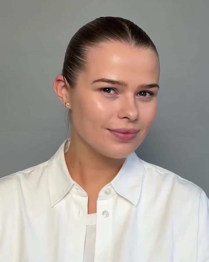 Portrait of a woman with slicked-back hair and a white collared shirt, smiling slightly at the camera against a gray background.