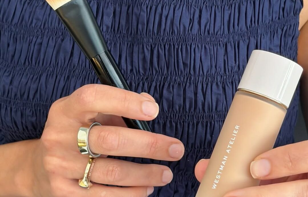 Person in a navy smocked top holds a makeup brush and a bottle of foundation.
