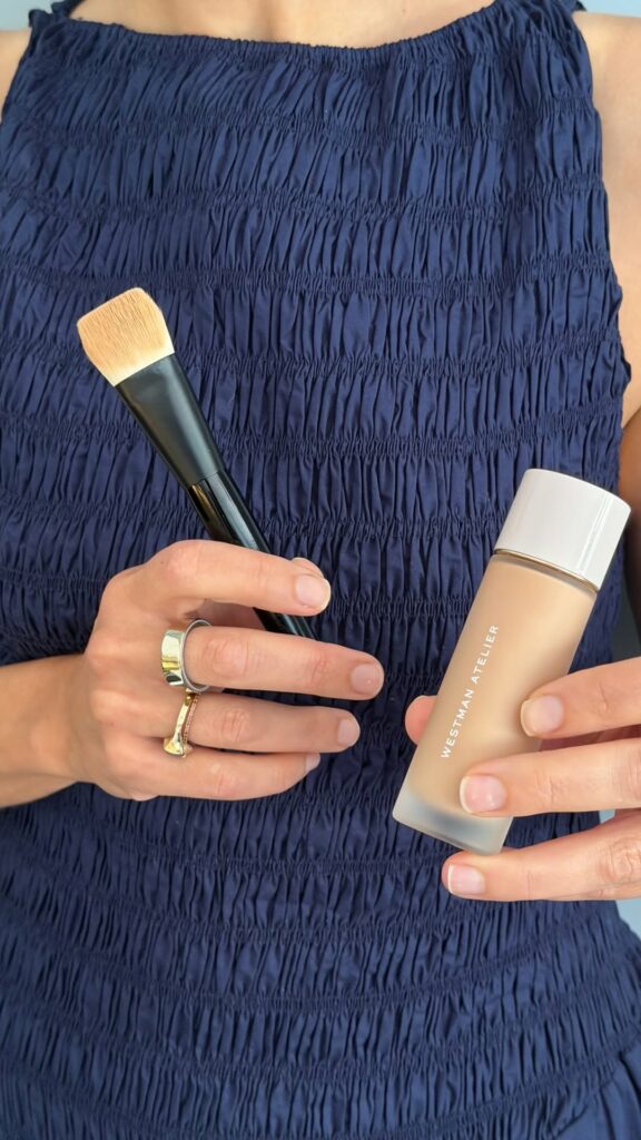 Person in a navy smocked top holds a makeup brush and a bottle of foundation.