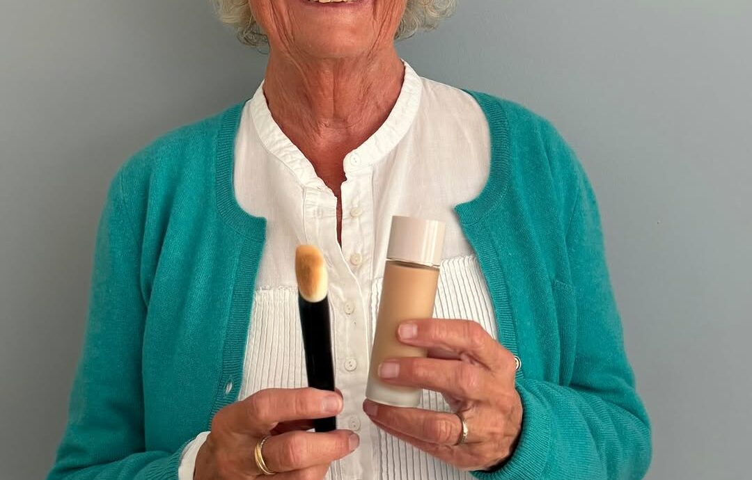 Smiling elderly woman with round glasses in a teal cardigan holds a makeup brush and a foundation bottle in front of a light gray wall.