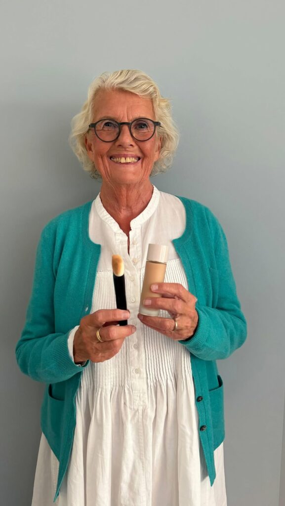 Smiling elderly woman with round glasses in a teal cardigan holds a makeup brush and a foundation bottle in front of a light gray wall.