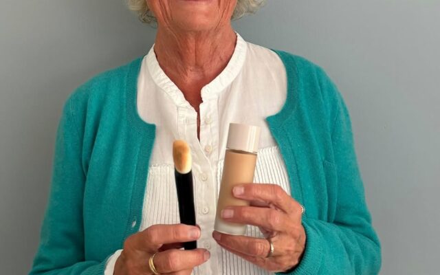 blogg-red-3962597710685558610-frame0 - Gimle Parfymeris blogg Smiling elderly woman with round glasses in a teal cardigan holds a makeup brush and a foundation bottle in front of a light gray wall.