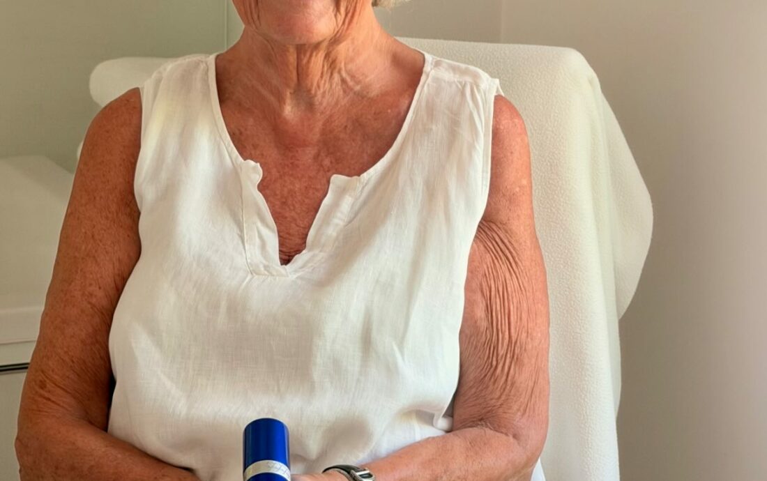 Smiling elderly woman with short white hair and round glasses sits in a white chair, holding a blue bottle (cosmetic product).