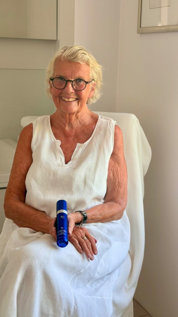 Smiling elderly woman with short white hair and round glasses sits in a white chair, holding a blue bottle (cosmetic product).