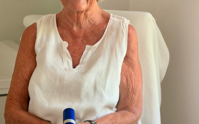 blogg-red-3967670596895098226-frame0 - Gimle Parfymeris blogg Smiling elderly woman with short white hair and round glasses sits in a white chair, holding a blue bottle (cosmetic product).