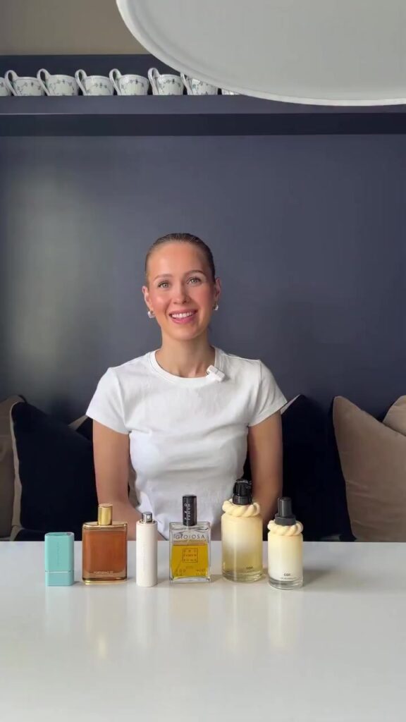 Smiling woman sits at a table with six perfume bottles lined up in front of her on a white surface.