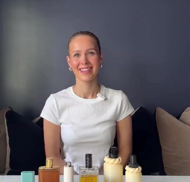 Smiling woman sits at a table with six perfume bottles lined up in front of her on a white surface.