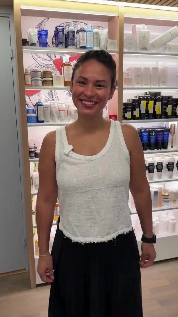 Smiling woman in a white sleeveless top standing in a skincare store with shelves of products behind her
