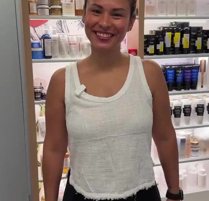 Smiling woman in a white sleeveless top standing in a skincare store with shelves of products behind her