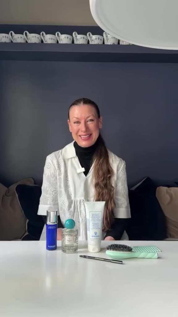 Smiling woman in a white embroidered top sits at a white table with skincare products: a blue bottle, clear perfume with a turquoise cap, a white tube, a mint brush, and a dark makeup pencil in front.