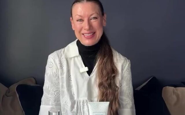 Smiling woman in a white embroidered top sits at a white table with skincare products: a blue bottle, clear perfume with a turquoise cap, a white tube, a mint brush, and a dark makeup pencil in front.