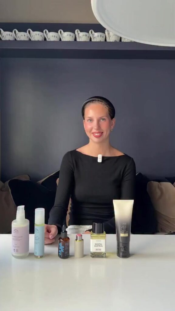 Smiling woman in a black top sits at a table with assorted skincare bottles and tubes in front of her.