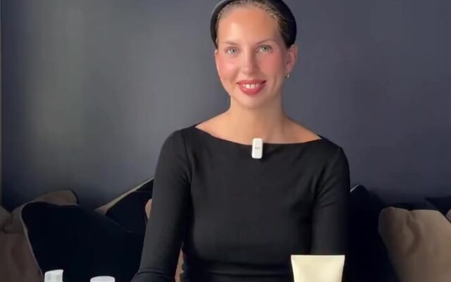 Smiling woman in a black top sits at a table with assorted skincare bottles and tubes in front of her.