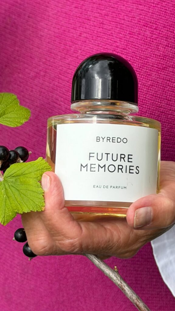 Perfume bottle labeled 'Byredo Future Memories' being held by a hand against a bright pink background with green leaves and dark berries nearby.
