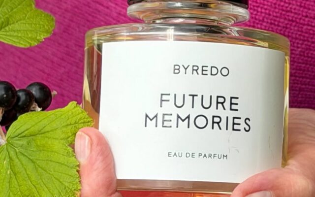 Perfume bottle labeled 'Byredo Future Memories' being held by a hand against a bright pink background with green leaves and dark berries nearby.