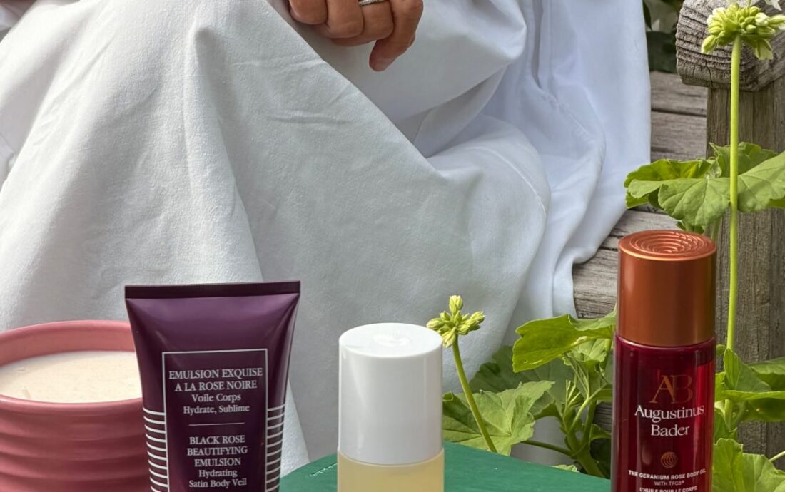 Person in a white outfit sits on a wooden bench, holding a white flower with green leaves; skincare products are arranged on a rustic table in front of them.