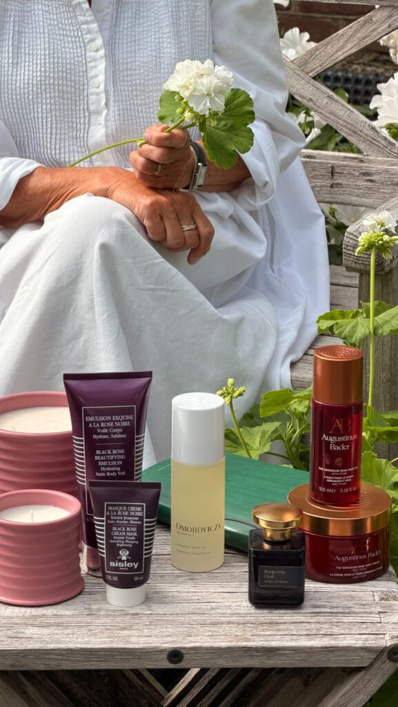 Person in a white outfit sits on a wooden bench, holding a white flower with green leaves; skincare products are arranged on a rustic table in front of them.