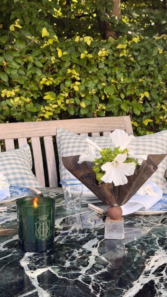 Outdoor dining scene with a metal flower centerpiece, white blooms, a green glass candle, and blue-check cushions against a leafy hedge.