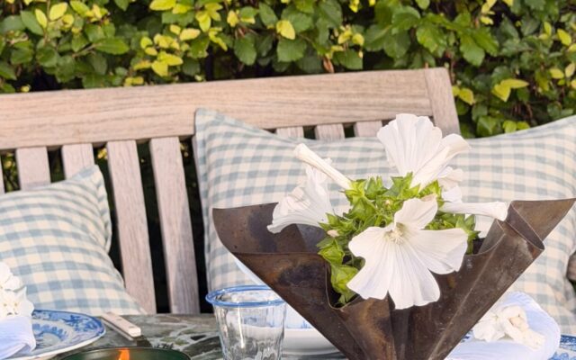 Outdoor dining scene with a metal flower centerpiece, white blooms, a green glass candle, and blue-check cushions against a leafy hedge.