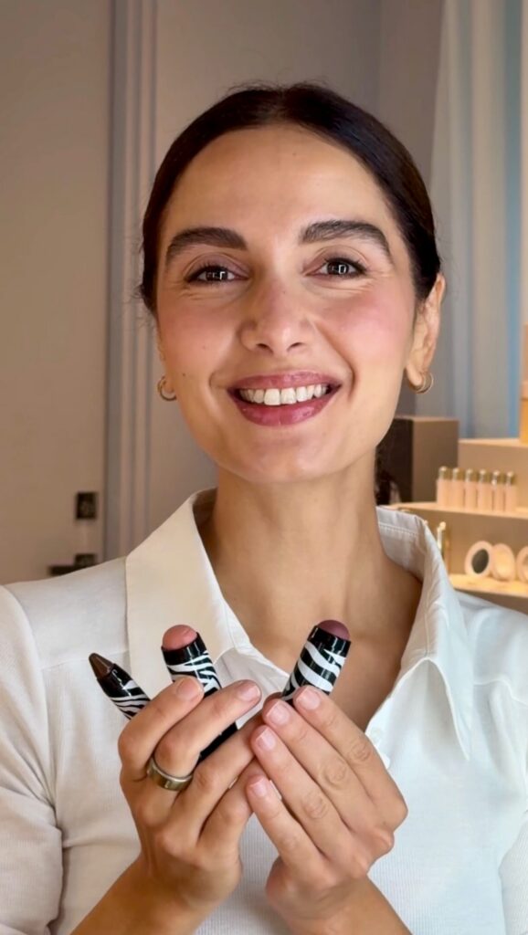 Smiling woman in a white shirt holds three zebra-striped lipstick tubes toward the camera in a cosmetics setting.