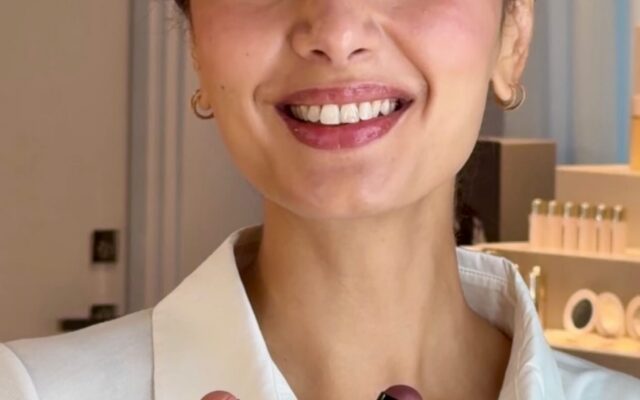 Smiling woman in a white shirt holds three zebra-striped lipstick tubes toward the camera in a cosmetics setting.