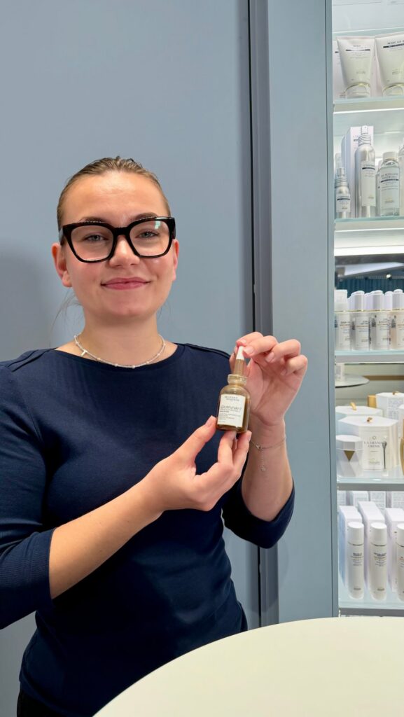 Smiling woman wearing black glasses holds a small bottle of serum in a skincare shop, with shelves of products in the background.