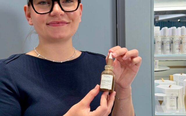Smiling woman wearing black glasses holds a small bottle of serum in a skincare shop, with shelves of products in the background.