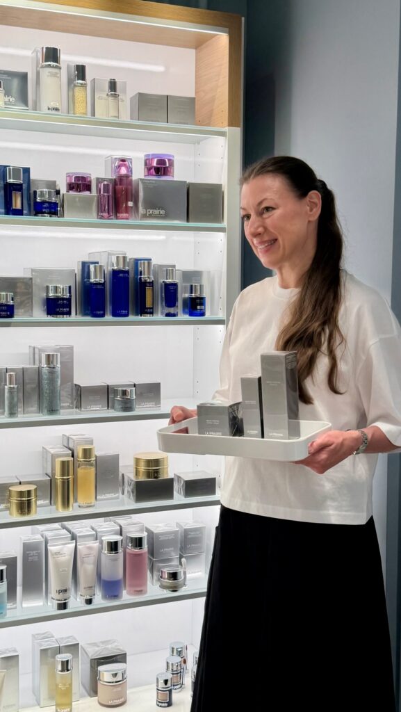 Smiling woman in a white top holds a tray of La Prairie skincare boxes in a bright store display with shelves of products behind her.