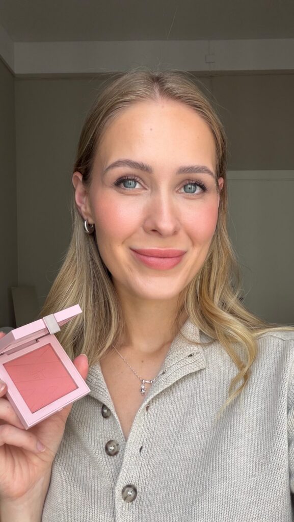 Close-up portrait of a smiling woman with blonde hair and blue eyes, holding a pink blush palette in front of her.