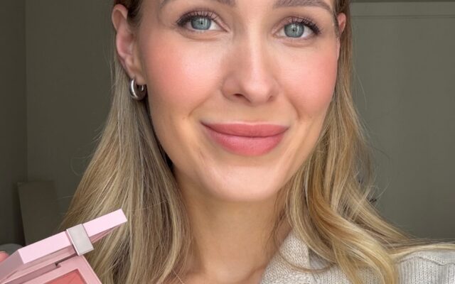 Close-up portrait of a smiling woman with blonde hair and blue eyes, holding a pink blush palette in front of her.
