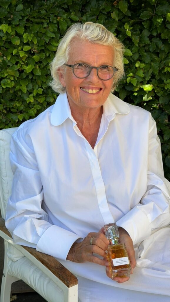 Smiling elderly woman with short gray hair and round glasses in a white shirt, sitting outdoors and holding a bottle of perfume.