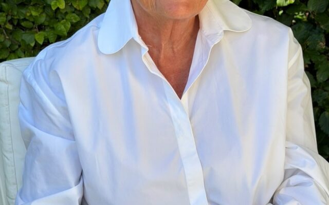 Smiling elderly woman with short gray hair and round glasses in a white shirt, sitting outdoors and holding a bottle of perfume.