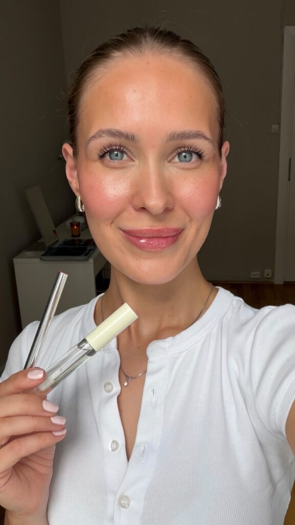 Smiling woman with blue eyes holds makeup tubes (lip gloss and mascara) near her face, wearing a white top.