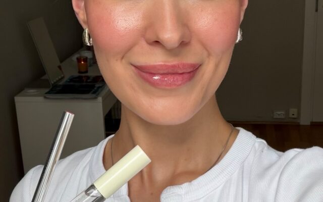 blogg-red-3983654524543453927-frame0 - Gimle Parfymeris blogg Smiling woman with blue eyes holds makeup tubes (lip gloss and mascara) near her face, wearing a white top.