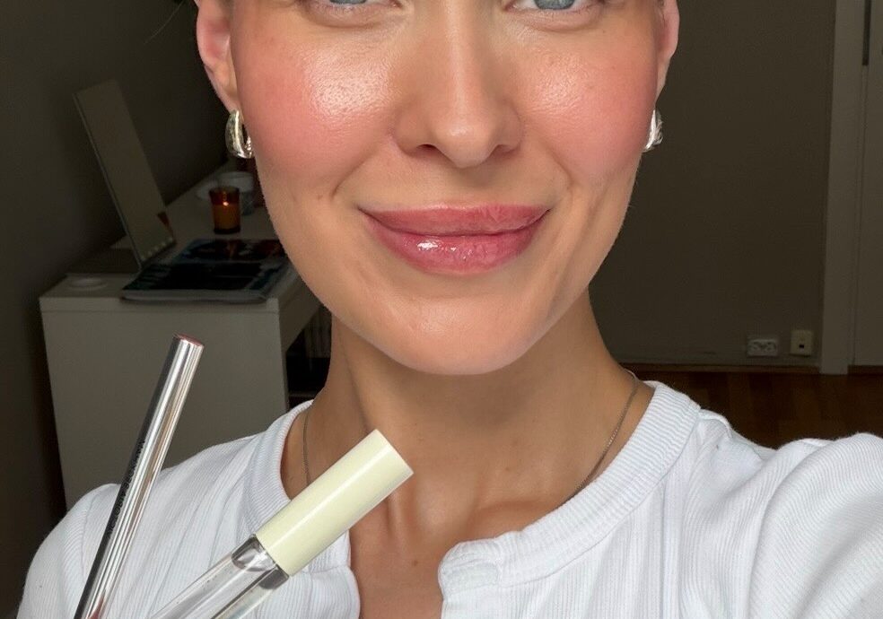 Smiling woman with blue eyes holds makeup tubes (lip gloss and mascara) near her face, wearing a white top.