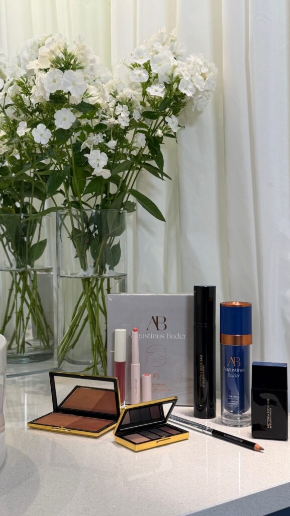 White flower arrangement in glass vases behind a cosmetics display on a countertop with lipsticks and palettes nearby.